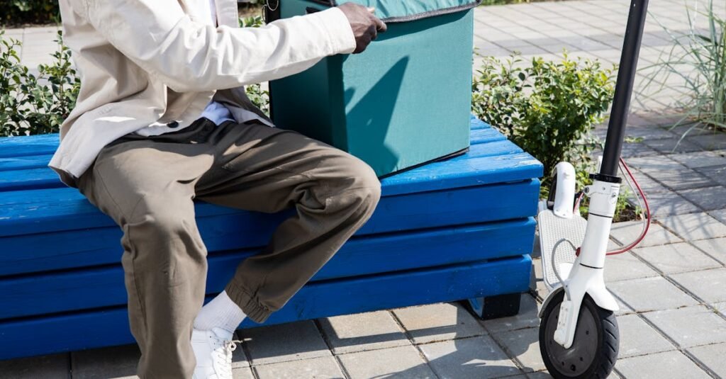 A delivery person with a thermal bag sitting on a blue bench next to an electric scooter.